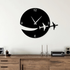 Puede incluir: Un reloj de pared negro con un diseño circular, con un recorte y dos siluetas de avión. Las manecillas del reloj son plateadas. El reloj está montado en una pared blanca sobre un mueble de madera con altavoces y objetos decorativos.