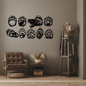 Puede incluir: Un conjunto de 12 decoraciones de pared de metal negro en forma de huellas de patas de oso. Las huellas de patas están recortadas en una variedad de diseños, incluyendo una cabeza de oso, una huella de pata de oso y una escena de bosque. Las huellas de patas están dispuestas en un patrón escalonado en una pared marrón claro.