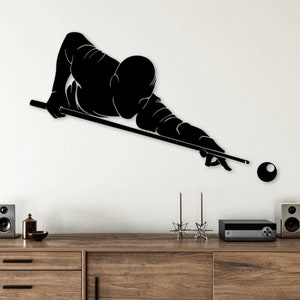 Puede incluir: Arte de pared en silueta negra de una persona jugando al billar, con un taco y una sola bola de billar. El arte está montado en una pared blanca sobre una cómoda de madera con un sistema estéreo y dos altavoces.