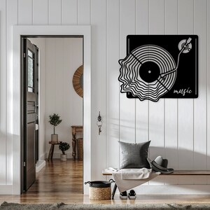 May include: Black metal wall art depicting a stylised record player with the word "music" beneath it. The art is mounted on a white wall above a wooden bench with a grey throw blanket and a brown woven basket.