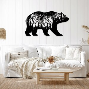 Puede incluir: Arte de pared de metal negro que representa la silueta de un oso con una cordillera y estrellas en su interior. El oso está colocado en una pared blanca, con un sofá blanco y una mesa de café en primer plano.