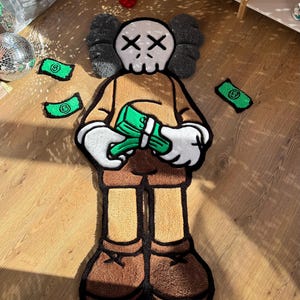 Puede incluir: Una alfombra de tufting con forma de personaje de dibujos animados con cabeza de calavera y ojos en forma de X, que viste una camisa beige y pantalones marrones. El personaje sostiene un fajo de dinero verde, con billetes verdes adicionales.