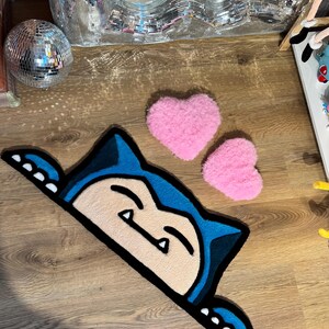 Puede incluir: Una alfombra con forma de personaje de dibujos animados azul y crema con un contorno negro, colocada sobre un suelo de madera. Dos alfombras rosas en forma de coraz&oacute;n tambi&eacute;n est&aacute;n en el suelo. Una bola de discoteca y un espejo decorativo est&aacute;n en el fondo.