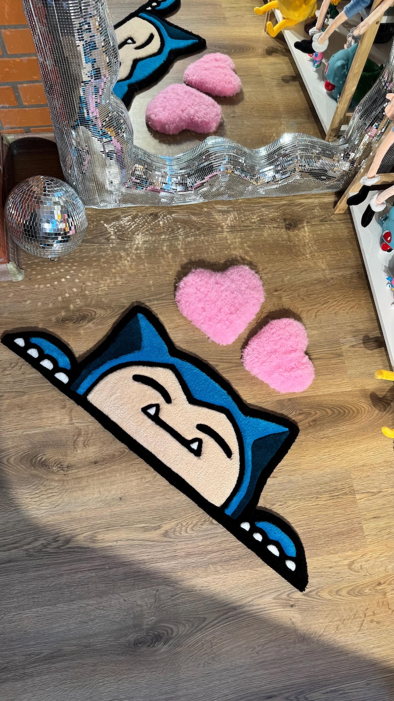 Puede incluir: Una alfombra con forma de personaje de dibujos animados azul y crema con un contorno negro, colocada sobre un suelo de madera. Dos alfombras rosas en forma de coraz&oacute;n tambi&eacute;n est&aacute;n en el suelo. Una bola de discoteca y un espejo decorativo est&aacute;n en el fondo.