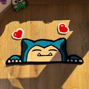 Handmade tufted rug showing kawaii sleepy character peeking with blue ears and soft paws, surrounded by cute red hearts. Plush fluffy pile creates cozy soft texture. Perfect as anime rug, kawaii rug, or tufted carpet for bedroom décor, nursery, or studio. Unique handmade rug gift for fans of cute rugs, couples, or friends. Adds cozy charm, playful vibe, and romantic cheerful accent to interiors