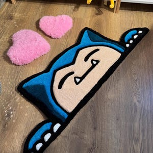 Puede incluir: Una alfombra azul, negra y beige con forma de personaje de dibujos animados, con dos alfombras rosas en forma de coraz&oacute;n. La alfombra del personaje tiene una cara sonriente y est&aacute; sobre un suelo de madera. Una adici&oacute;n divertida y l&uacute;dica a cualquier habitaci&oacute;n.