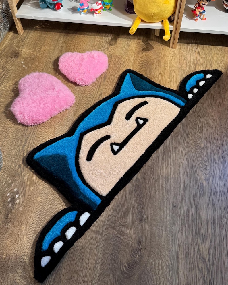 Puede incluir: Una alfombra azul, negra y beige con forma de personaje de dibujos animados, con dos alfombras rosas en forma de coraz&oacute;n. La alfombra del personaje tiene una cara sonriente y est&aacute; sobre un suelo de madera. Una adici&oacute;n divertida y l&uacute;dica a cualquier habitaci&oacute;n.