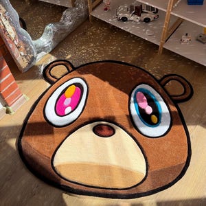 Pode incluir: Tapete em forma de urso marrom com um design de desenho animado. O tapete apresenta um focinho marrom claro, olhos grandes com detalhes em rosa, amarelo e azul e contornos pretos. O tapete está colocado em um piso de madeira.