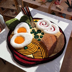 Könnte beinhalten: Ein getufteter Teppich in Form einer Ramenschüssel mit gelben Nudeln, zwei weiß-orangen gekochten Eiern, grünen Algen, braunem Fleisch, weiß-roten Fischkuchen und einem Paar brauner Essstäbchen, alles mit einem dicken schwarzen Rand.