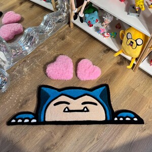 Puede incluir: Una alfombra Snorlax azul y beige con un contorno negro y dos alfombras rosas en forma de coraz&oacute;n. La alfombra est&aacute; sobre un suelo de madera. Estanter&iacute;as en el fondo exhiben peluches de personajes de dibujos animados.