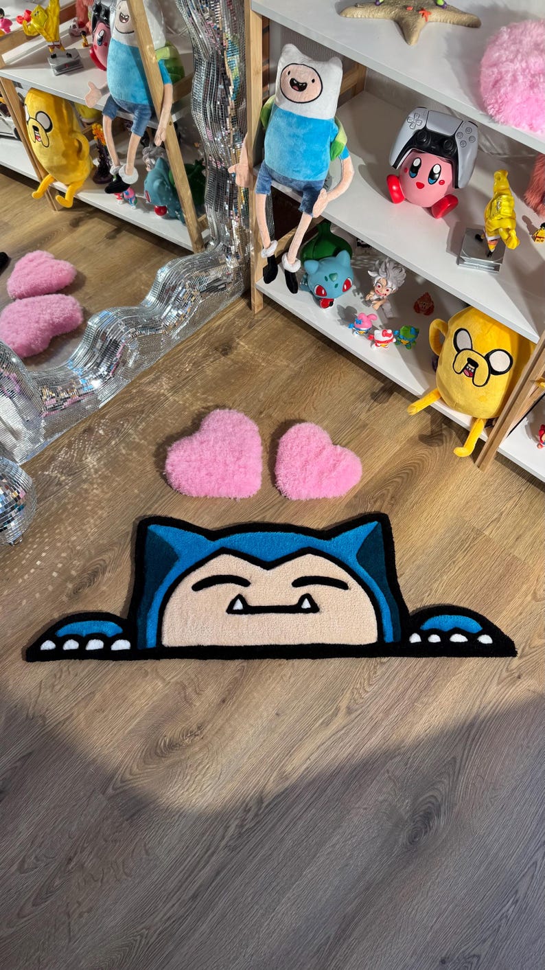 Puede incluir: Una alfombra Snorlax azul y beige con un contorno negro y dos alfombras rosas en forma de coraz&oacute;n. La alfombra est&aacute; sobre un suelo de madera. Estanter&iacute;as en el fondo exhiben peluches de personajes de dibujos animados.