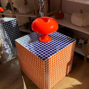 Pode incluir: Uma mesa lateral em forma de cubo coberta com uma grade de pequenos azulejos quadrados laranja e azul escuro. A superfície superior apresenta uma borda de azulejos azul escuro em torno de uma área central laranja. Uma lâmpada laranja brilhante em forma de cogumelo está sobre a mesa.