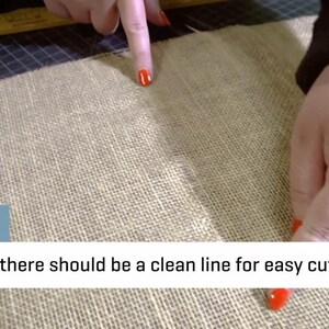 May include: Close-up of a beige burlap fabric with a visible seam. The text "TIP: Now there should be a clean line for easy cutting" is displayed in a white box. A person's fingers, with red nail polish, point to the seam.