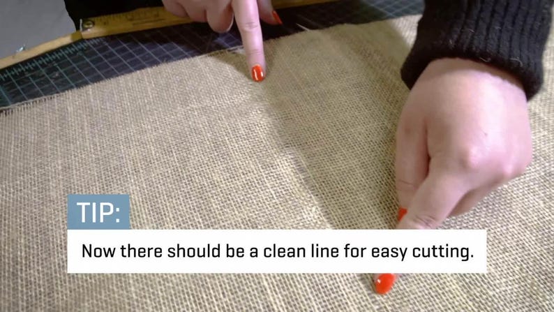May include: Close-up of a beige burlap fabric with a visible seam. The text "TIP: Now there should be a clean line for easy cutting" is displayed in a white box. A person's fingers, with red nail polish, point to the seam.