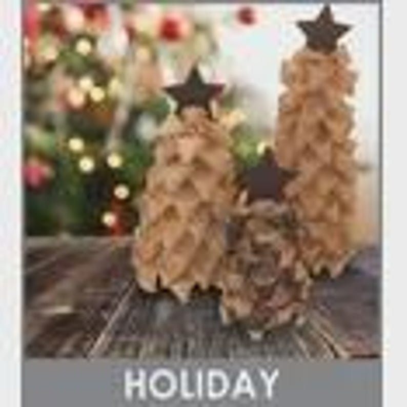 May include: Three handmade Christmas trees of varying heights, crafted from natural materials, stand on a wooden surface. Each tree is adorned with a dark brown star at the top. The background features a blurred Christmas tree with twinkling lights, evoking a festive holiday atmosphere.