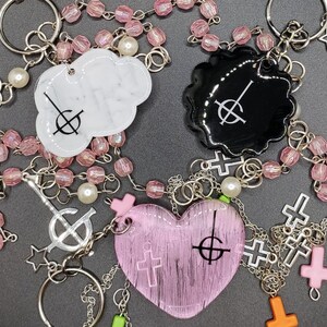 May include: A pink heart-shaped charm with a black symbol, a white cloud-shaped charm with a black symbol, and a black circle charm with a white symbol. All charms are attached to a silver chain with pink beads and silver crosses.