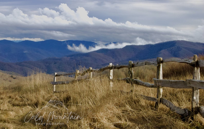 K&ouml;nnte beinhalten: Ein verwitterter Holzzaun erstreckt sich &uuml;ber ein grasbewachsenes Feld mit Blick auf ferne blaue Berge und fluffige wei&szlig;e Wolken am Himmel.