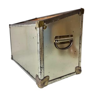 Op de afbeelding: Een vintage metalen opbergdoos met een zilveren afwerking en hoekbeschermers in messingkleur. De doos heeft een handvat aan &eacute;&eacute;n kant en is licht schuin. De doos wordt waarschijnlijk gebruikt voor het opbergen van items.