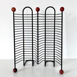 May include: A black metal wire rack featuring a zigzag design with multiple horizontal slots for storage. The structure is supported by vertical rods topped and bottomed with small, round, reddish-brown wooden spheres at each corner and center.