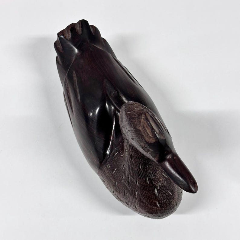 May include: A dark brown, carved wooden duck figurine. The duck is depicted with its head tucked down, and the wood has a glossy finish. The carving shows detailed feather textures on the body and wings. The figurine is set against a white background.