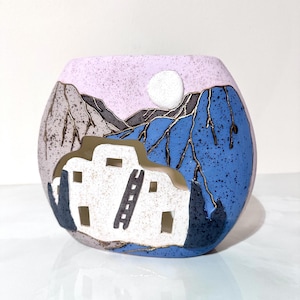 May include: A ceramic vase with a Southwestern design. The vase features a white pueblo-style building with a ladder, set against a backdrop of blue and gray mountains, a pink sky, and a white moon. The vase has a speckled texture.