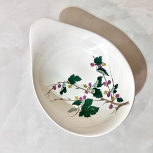 May include: A white, oval-shaped serving dish with a decorative botanical design. The design features a branch with green leaves, purple berries, and yellow buds. The dish has a smooth, glossy finish and a curved handle.