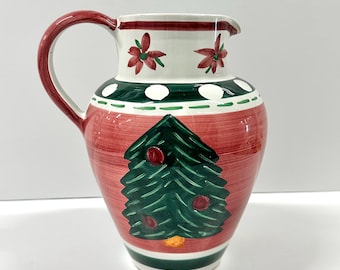 May include: A ceramic Christmas pitcher with a red body and a green and white rim. The pitcher features a green Christmas tree design with red ornaments and a red handle. The top rim has red floral accents.
