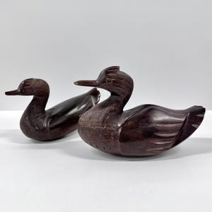 May include: Two hand-carved wooden duck figurines. The ducks are a dark brown color with detailed carving, including feathers and beaks. One duck is in front of the other, both facing left. The ducks are on a white surface.
