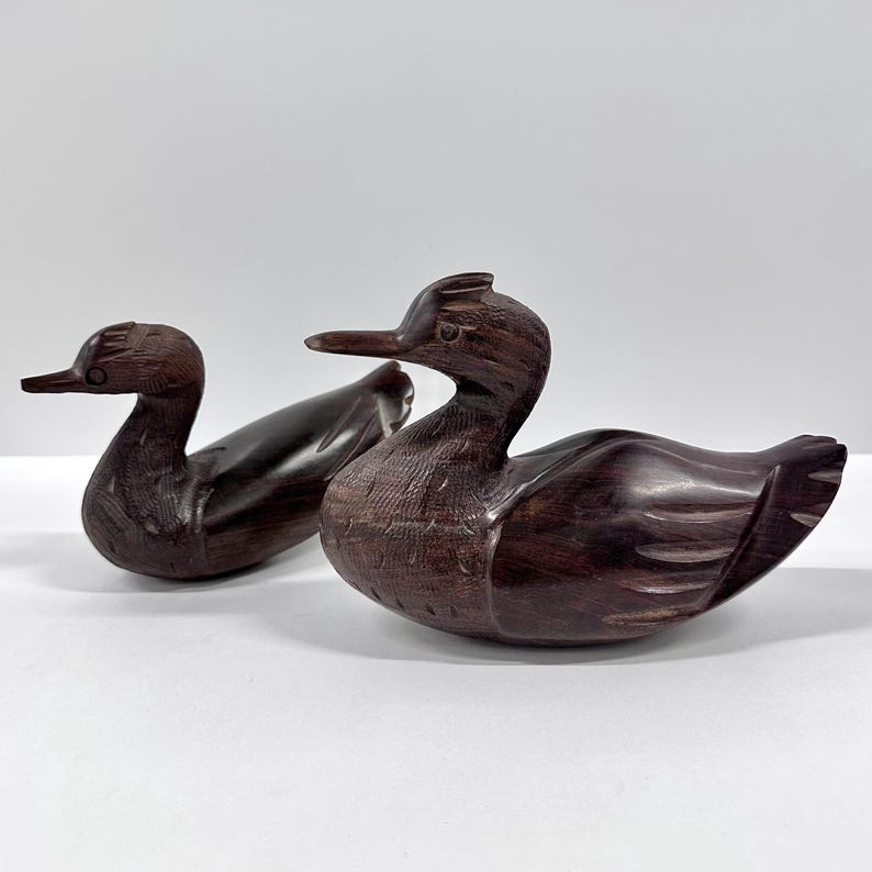 May include: Two hand-carved wooden duck figurines. The ducks are a dark brown color with detailed carving, including feathers and beaks. One duck is in front of the other, both facing left. The ducks are on a white surface.