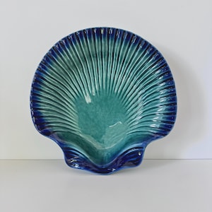 May include: A ceramic dish shaped like a large scallop shell, featuring a glossy glaze that transitions from a deep navy blue at the outer edges to a vibrant turquoise green in the center, with prominent raised ridges radiating outward.