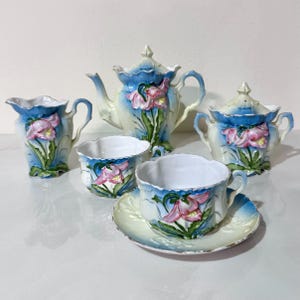 May include: A vintage porcelain tea set featuring a teapot, sugar bowl, creamer, two teacups, and a saucer. The set is painted with a floral design in pink, blue, and green. The teapot has a curved spout and a lid with a finial.