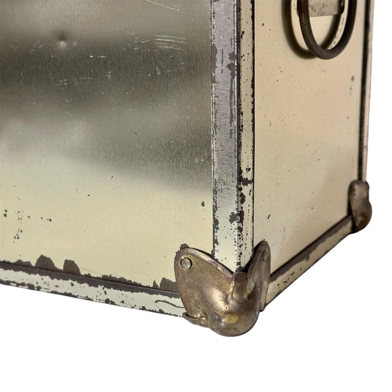 Op de afbeelding: Close-up van een vintage metalen koffer met een verweerde, verouderde uitstraling. De koffer heeft een lichtgele afwerking met zichtbare slijtage, waaronder krassen en afgebladderde verf. Metalen hoekbeschermers en een handvat zijn zichtbaar.