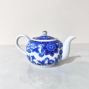 May include: A white porcelain teapot with a blue floral pattern. The teapot has a curved handle, a spout, and a lid with a gold-colored knob. The floral design is a classic blue and white pattern.