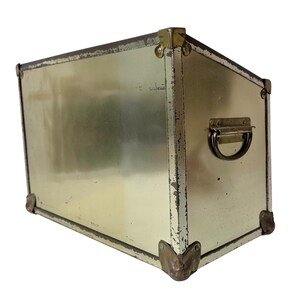 Op de afbeelding: Een rechthoekige, vintage metalen kist met een lichtgouden afwerking. De kist heeft een handvat aan de zijkant en hoekbeschermers in messingkleur. De randen vertonen tekenen van slijtage, wat het een rustieke uitstraling geeft.