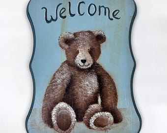 May include: Wooden sign with a light blue background and a brown teddy bear illustration. The word "Welcome" is written in cursive at the top. The sign has a decorative, wavy border.