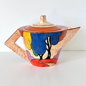 May include: A ceramic teapot featuring a speckled beige base with a colorful abstract landscape design. The teapot has a geometric handle, a pointed spout, and a matching lid. The surface displays bold shapes in blue, yellow, red, and purple.