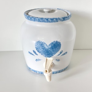 May include: A white ceramic beverage dispenser featuring a blue sponge-painted heart design on the front. The container includes a matching lid and a white plastic spigot at the base. The rim and base are decorated with a blue speckled pattern.