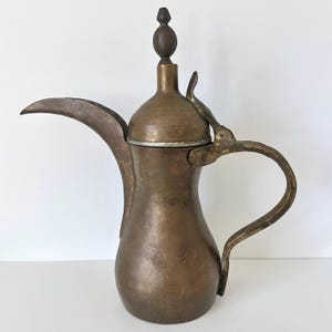 May include: A tall, weathered brass coffee pot featuring a long, curved spout, a rounded body, and a slender handle. The lid is topped with a decorative finial and includes a hinged mechanism that connects to the handle, showing a patina.