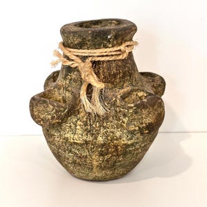 May include: A textured, dark brown and green multi-opening vase with a weathered, earthy finish. The vessel features a central opening at the top and several smaller, rounded openings around its body, all tied with a piece of natural jute twine.