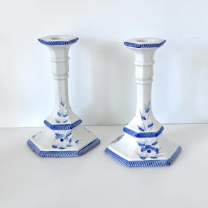May include: A pair of white ceramic candle holders featuring a hexagonal base and a flared top. Each piece is decorated with hand-painted blue floral motifs and blue rope-style trim along the edges of the base, the mid-section, and the top rim.