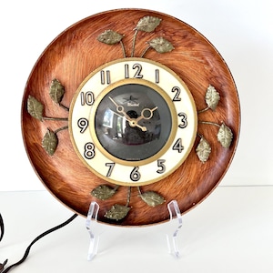 May include: A round wall clock featuring a wood-grain finish background with metallic gold-toned leaf accents. The center dial has a cream-colored face with black numerals, a gold-toned frame, and the text United printed above the clock hands.