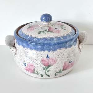 May include: A round ceramic casserole dish with a lid, featuring a speckled cream base, a blue rim, and hand-painted pink flowers with green stems. The pot includes two side handles and a small round knob on the lid, all with a speckled finish.
