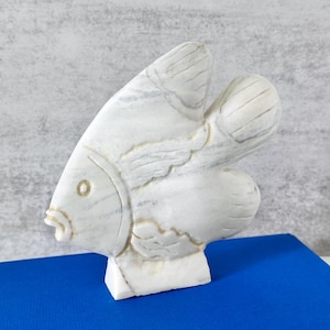 May include: A white marble sculpture of a fish with gray veining. The fish is depicted in profile with carved details for the eye, mouth, fins, and tail. The sculpture is mounted on a rectangular base and features a smooth, polished stone surface.