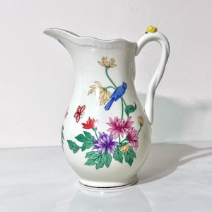 May include: White ceramic pitcher with a floral design featuring a blue bird, butterflies, and colorful flowers. The pitcher has a curved handle and a decorative rim. The base has a gold trim.