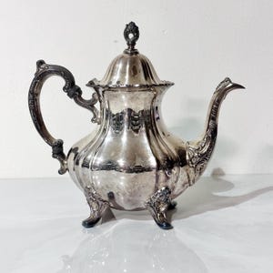 May include: A silver-colored teapot with ornate detailing. The teapot has a curved handle, a spout, and a lid with a decorative finial. The teapot sits on three small feet. The design is vintage and elegant.