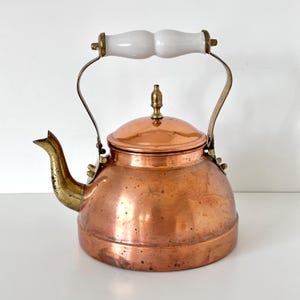 May include: A copper tea kettle featuring a rounded body, a brass spout, and a matching brass lid with a small finial. The kettle has a tall, arched metal handle topped with a smooth, white, contoured ceramic grip for handling the vessel.