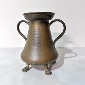 May include: A vintage bronze-toned metal vase with two curved handles and a wide, shallow top. The vase has a bulbous body and rests on three claw-shaped feet. The metal has a hammered texture, giving it a rustic appearance.