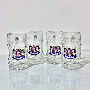 May include: Four clear glass beer mugs with handles. Each mug has a decorative design with the words "Kulmbacher Mönchshof" in blue and red. The mugs are cylindrical with a textured pattern and a small handle.