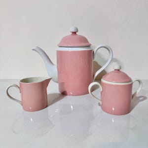 May include: A three-piece tea set in a soft pink color with white accents. The set includes a teapot, a creamer, and a sugar bowl. The teapot has a lid and a curved spout. The creamer and sugar bowl have handles.