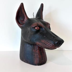 May include: A sculpted wax candle shaped like a Doberman dog head. The candle features a black coat with deep red highlights on the ears, muzzle, and neck. It has pointed ears, a long snout, and distinct red-colored eyes with a visible wick on top.